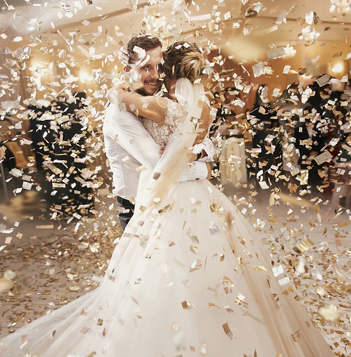 Gorgeous bride and stylish groom dancing under golden confetti at wedding reception. Happy wedding couple performing first dance in restaurant. Romantic moments